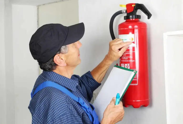 A professional technician performing a safety check on a wall-mounted fire extinguisher while taking notes on a clipboard.