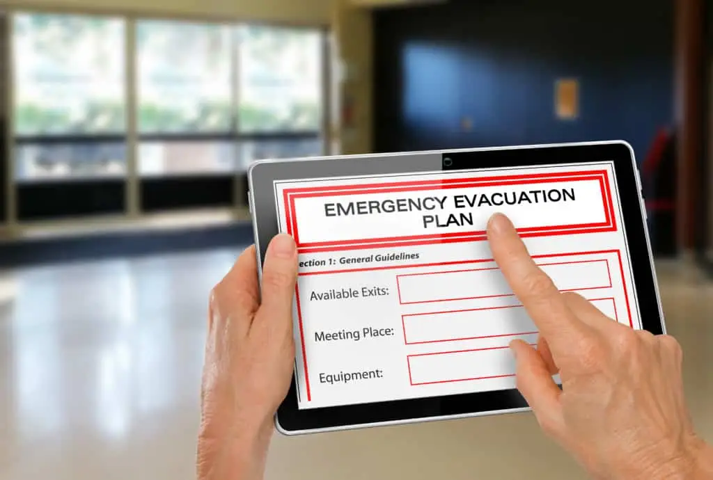 A person holds a tablet displaying an emergency evacuation plan with sections for general guidelines, available exits, meeting place, and equipment, indicating preparedness for an emergency situation.