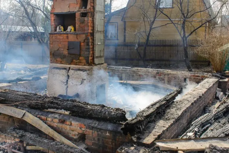 The remnants of a house after a fire, with only a chimney and parts of the structure still standing amidst the ruins and smoke.