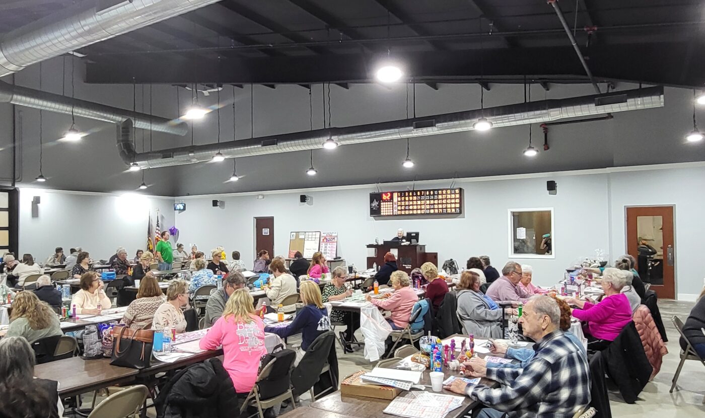 Large group of people sitting at tables in a spacious hall, playing bingo. Bingo board is visible on the wall at the front, with a person calling numbers. The atmosphere appears lively and social.