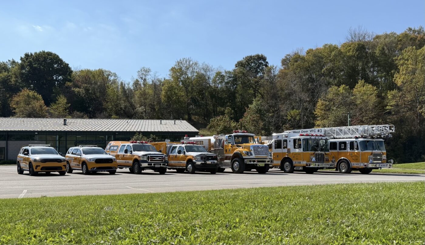 A row of West Deer Volunteer Fire Company emergency vehicles, including two SUVs, two pickup trucks, and two fire engines, parked in a paved lot in front of a building.