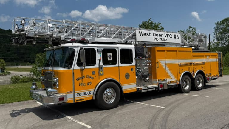A yellow fire truck with an extended ladder labeled “West Deer VFC #3 290 TRUCK” is parked in a lot on a sunny day with trees and blue sky in the background.