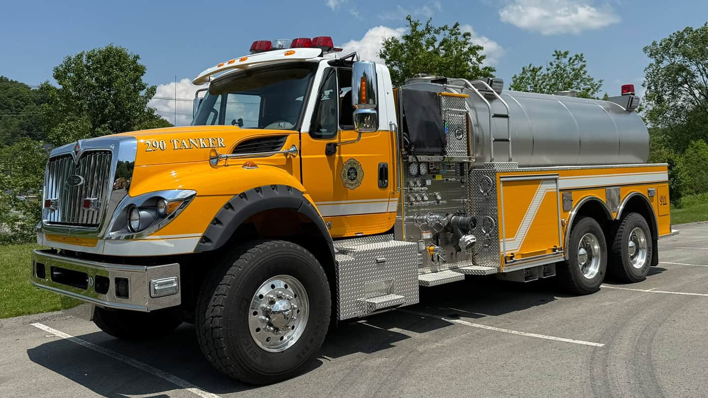 A yellow fire tanker truck with chrome details is parked in a sunny parking lot. Trees and blue sky are visible in the background.