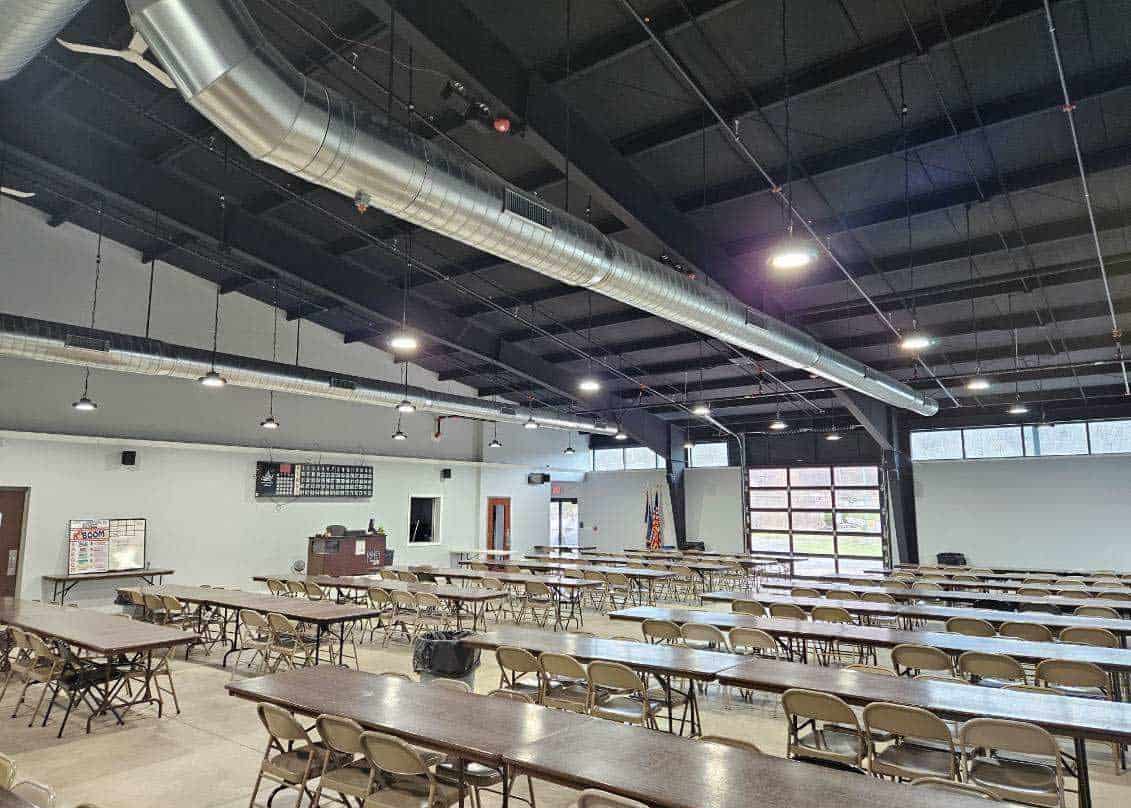 Large hall with rows of empty tables and chairs, high ceiling with exposed ducts, podium at the front, and windows along one wall.