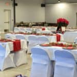 Room set up for a holiday event with tables covered in white cloths, red runners, festive decorations, and poinsettias as centerpieces. White chairs surround the tables in a brightly lit hall.