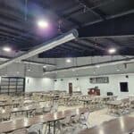 Large, empty cafeteria with rows of tables and chairs under industrial-style lighting and exposed ductwork. A small stage or podium is visible at the far end of the room.