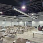 Large room with rows of empty tables and folding chairs, concrete floor, high ceiling with exposed ducts and lights, and a table with a register at the front near a garage-style door.