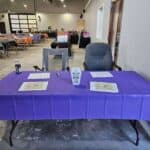A table with a purple tablecloth holds papers, a water bottle, and a small sign. Empty chairs sit behind it, and rows of tables and chairs are set up in the background, suggesting an event setup.