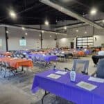 A large event hall with rows of tables covered in orange and purple tablecloths, folding chairs, papers, and decorations, prepared for a gathering or meeting.