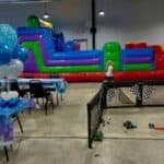 A child stands near a colorful inflatable obstacle course at an indoor party with decorated tables, chairs, balloons, and small toys scattered on the floor.