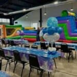 A decorated indoor party area with tables covered in blue tablecloths, balloon centerpieces, and a large colorful inflatable obstacle course in the background.