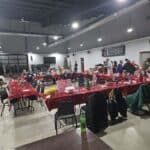 A large room with people standing and sitting at tables covered in red tablecloths, scattered with food, drinks, and gifts. Some chairs have coats draped over them. A decorated tree is visible in the background.