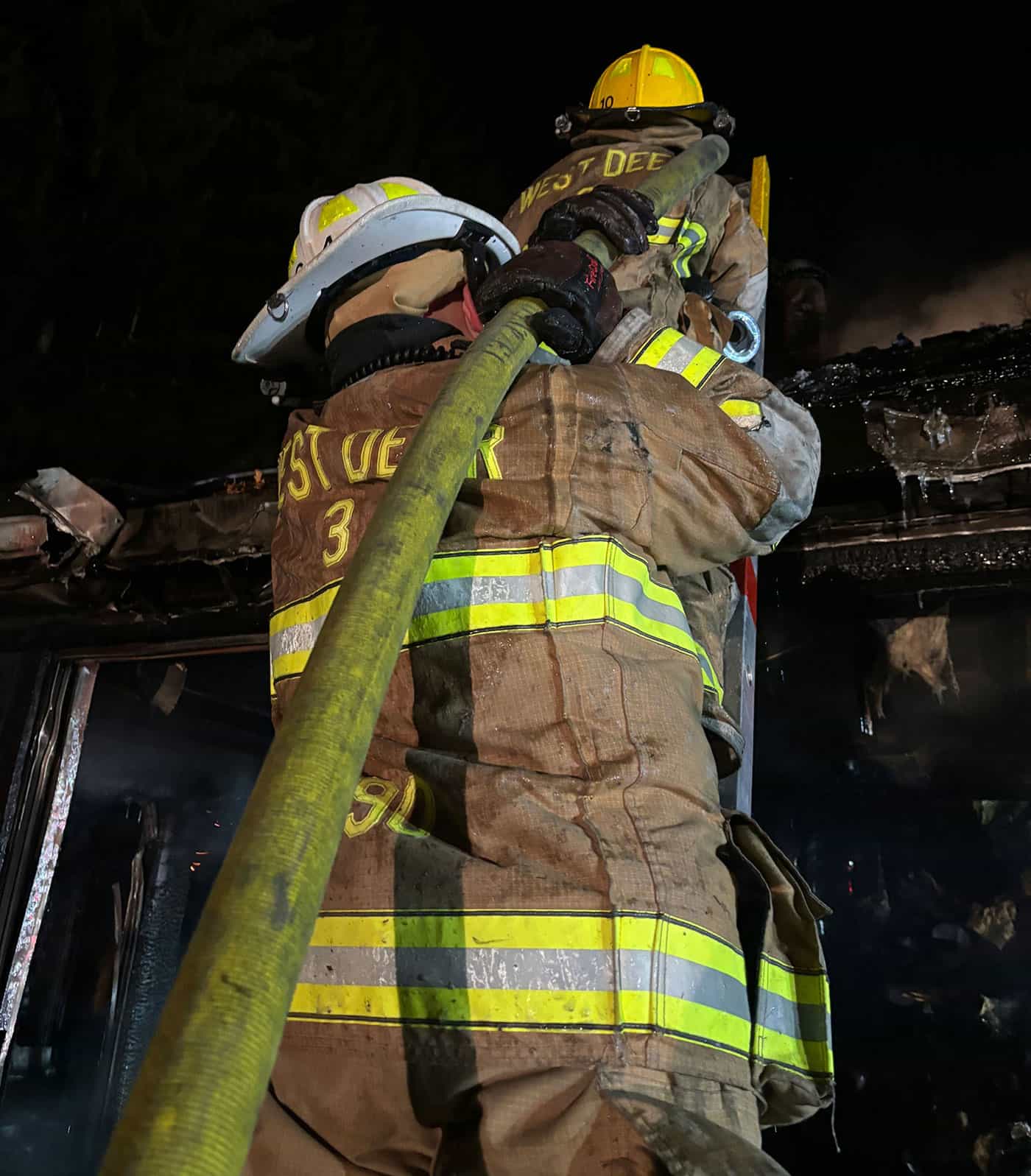 Two firefighters in full gear, one wearing a yellow helmet, work together holding a large hose in front of a burnt, charred building at night. Their uniforms read “WEST DEER.”.