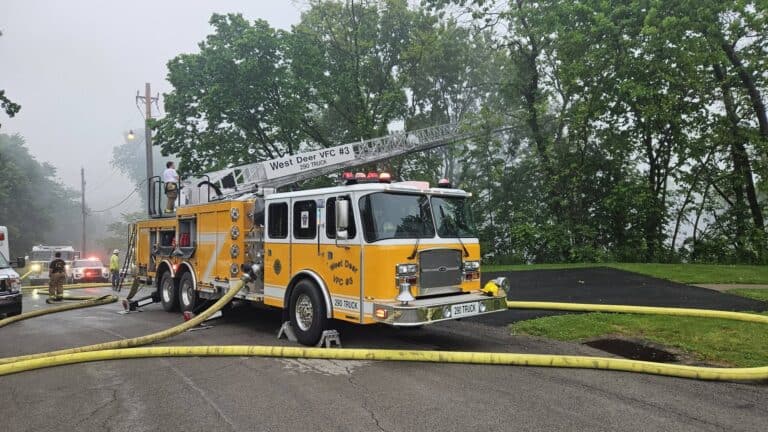 A yellow and white West Deer VFC ladder truck parked on a road with fire hoses deployed during an emergency response.