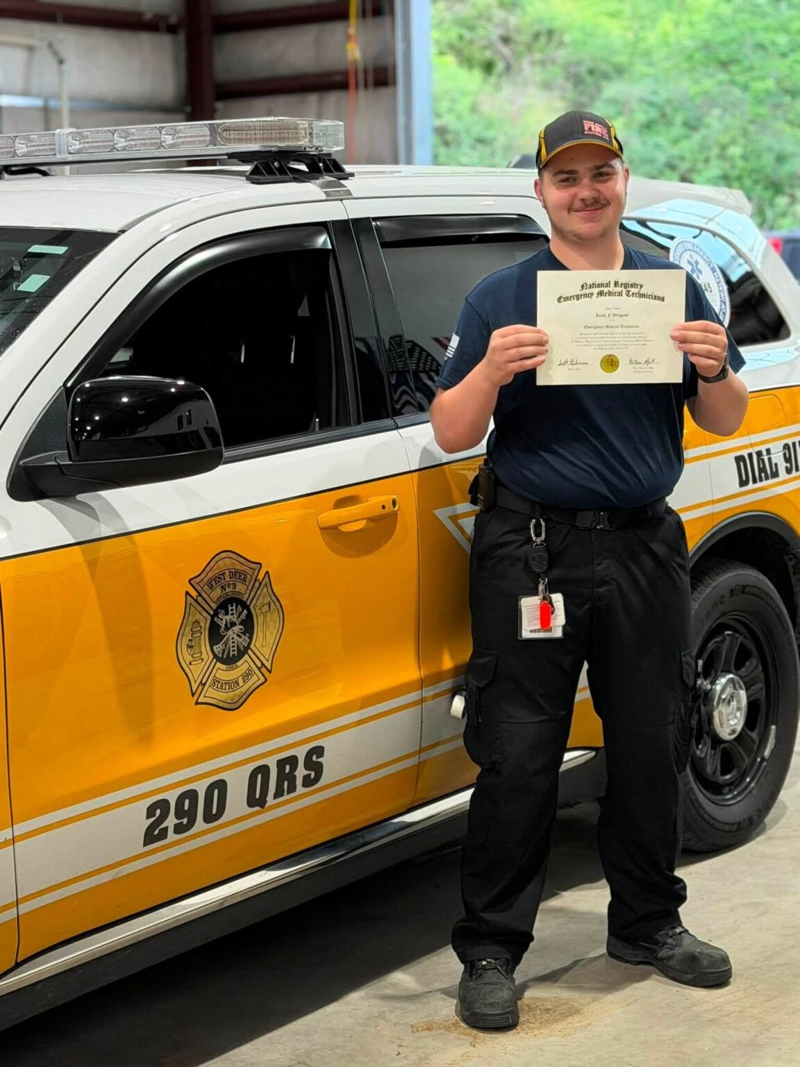 A smiling emergency medical technician in a uniform holding his NREMT certification in front of a West Deer emergency response vehicle.