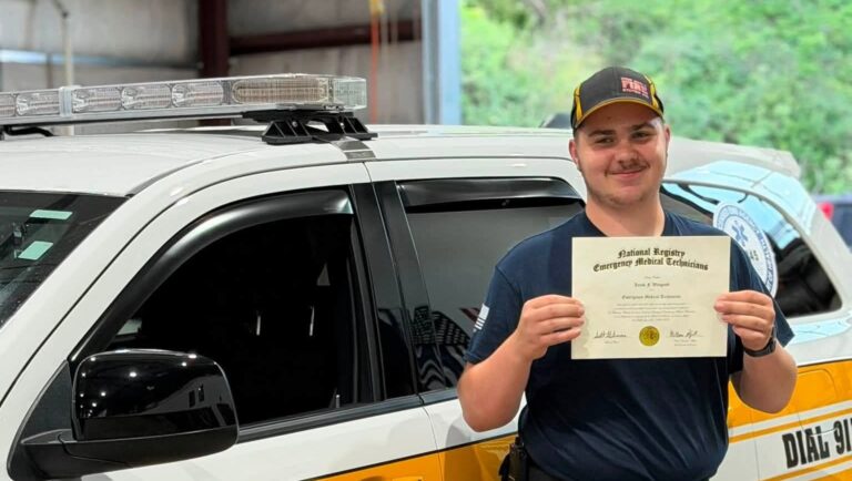 A smiling emergency medical technician in a uniform holding his NREMT certification in front of a West Deer emergency response vehicle.